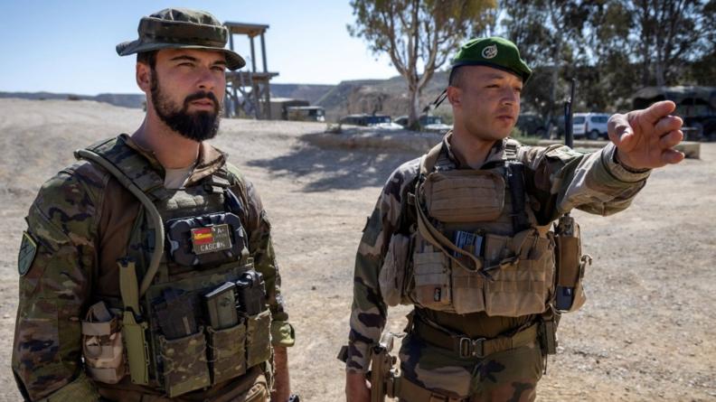 Deux soldats debout en conversation dans une zone aride, l’un désignant une direction.